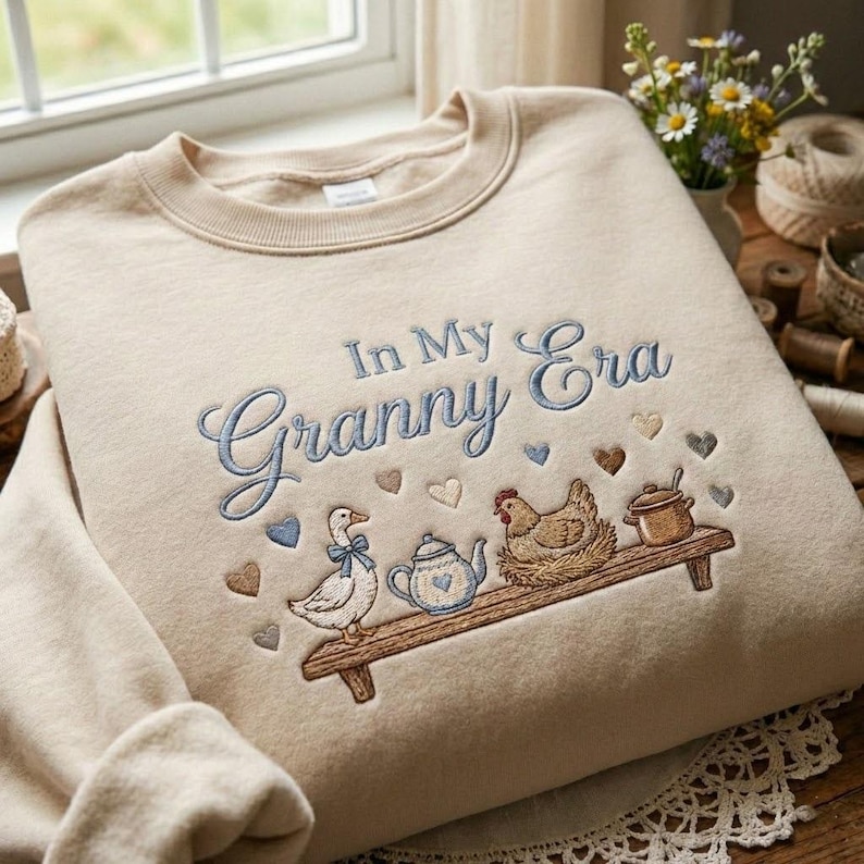 May include: Beige sweatshirt with the embroidered phrase "In My Granny Era" in blue script. The design includes a duck, teapot, chicken, and pot on a wooden shelf, with heart accents.