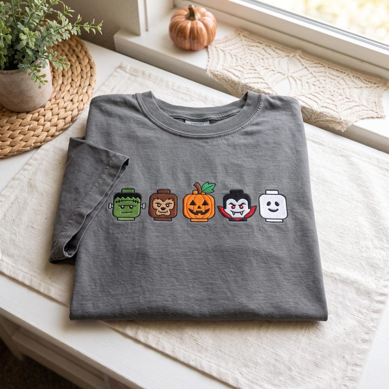 May include: A charcoal gray t-shirt featuring an embroidered row of five Halloween-themed character heads, including a green monster, a brown werewolf, an orange jack-o'-lantern, a vampire with fangs, and a plain white ghost-like figure.