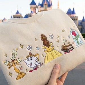 May include: Beige sweatshirt featuring embroidered Beauty and the Beast characters, including Belle in a yellow gown, Lumiere, Mrs. Potts, and Cogsworth. The design also includes a rose in a glass case, flowers, and stars.