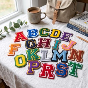 May include: A collection of twenty-one embroidered varsity-style letter patches in various colours including red, blue, green, yellow, purple, orange, and grey. Each letter features a bold block font with a contrasting border, arranged on a white surface.
