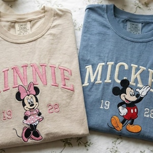 May include: Two Comfort Colors crewneck sweatshirts. One is tan with pink embroidered "MINNIE" and a Minnie Mouse graphic. The other is blue with white embroidered "MICKEY" and a Mickey Mouse graphic. Both have the numbers "19" and "28".