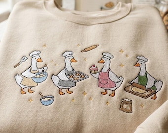 May include: A beige crewneck sweatshirt featuring an embroidered design of four white geese wearing chef hats and aprons. The geese are depicted baking with items including a whisk, a tray of cookies, a cupcake, a rolling pin, and a bag of flour.