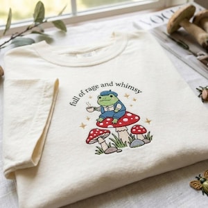 May include: An off-white t-shirt featuring an embroidered design of a green frog wearing a blue beret and overalls, sitting on a red mushroom while holding a cup. The text "full of rage and whimsy" is arched above the frog with small stars.