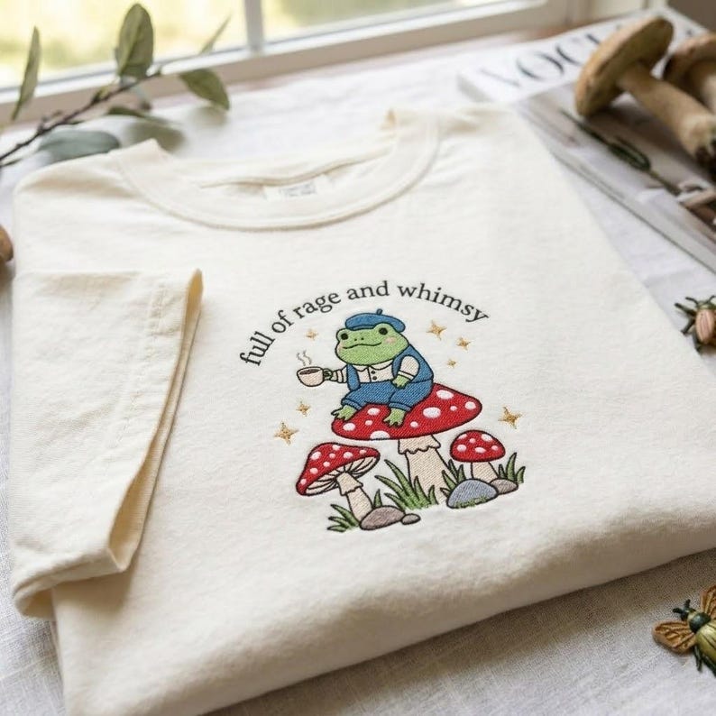 May include: An off-white t-shirt featuring an embroidered design of a green frog wearing a blue beret and overalls, sitting on a red mushroom while holding a cup. The text "full of rage and whimsy" is arched above the frog with small stars.