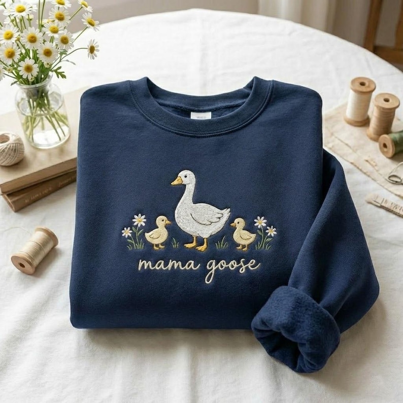 May include: Navy blue sweatshirt with an embroidered design featuring a white goose, two yellow ducklings, and the words "mama goose." The design is surrounded by embroidered flowers. The sweatshirt has a crew neck and long sleeves.