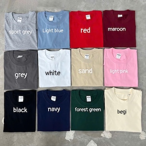May include: A collection of twelve crew neck t-shirts arranged in a grid, featuring various solid colors including sport grey, light blue, red, maroon, grey, white, sand, light pink, black, navy, forest green, and begi, each with a white label.