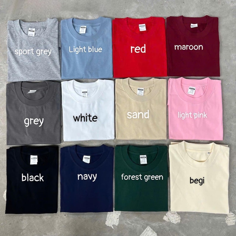 May include: A collection of twelve crew neck t-shirts arranged in a grid, featuring various solid colors including sport grey, light blue, red, maroon, grey, white, sand, light pink, black, navy, forest green, and begi, each with a white label.