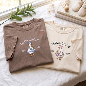 May include: A brown t-shirt and a cream t-shirt featuring a graphic of a goose carrying a gosling. The brown shirt reads DADA GOOSE EST. 2027 with a goose in a blue cap. The cream shirt reads MAMA GOOSE EST. 2027 with a goose in a pink bow.