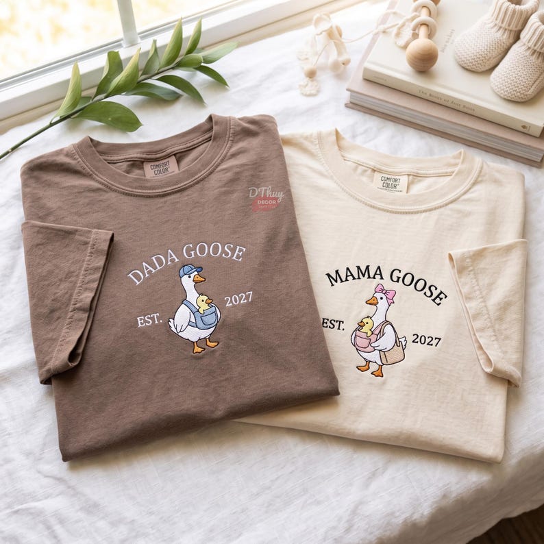 May include: A brown t-shirt and a cream t-shirt featuring a graphic of a goose carrying a gosling. The brown shirt reads DADA GOOSE EST. 2027 with a goose in a blue cap. The cream shirt reads MAMA GOOSE EST. 2027 with a goose in a pink bow.