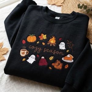 Pode incluir: Uma camisola preta de gola redonda com bordados coloridos de temas de outono, incluindo uma ab&oacute;bora, fogueira, fantasmas, uma caneca, um gorro, botas e uma fatia de tarte, com o texto "cozy season" escrito em letra cursiva castanha.