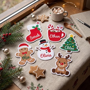Könnte beinhalten: Eine Sammlung von sechs personalisierten Weihnachtsanhängern, bestehend aus einem roten Strumpf mit der Aufschrift Sarah, einer roten Tasse mit Zuckerstangen mit der Aufschrift Ethan, einem Lebkuchenmann mit der Aufschrift Mia, einem Schneemann mit der Aufschrift Olivia, einem Baum mit der Aufschrift Chloe und einem Rentier mit der Aufschrift Noah.