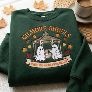 May include: A dark green crewneck sweatshirt featuring an embroidered design of a gazebo with two white ghosts holding a coffee cup. The text reads GILMORE GHOULS and WHERE YOU HAUNT, I WILL FOLLOW, surrounded by small orange, yellow, and red autumn leaves.