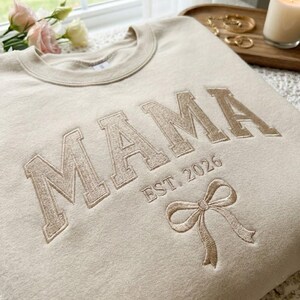 Peut inclure: Un sweat-shirt beige avec le mot "MAMA" brod&eacute; en police serif. En dessous, "EST. 2026" et un n&oelig;ud. Le sweat-shirt est sur une surface claire, avec des fleurs et des bijoux en arri&egrave;re-plan.