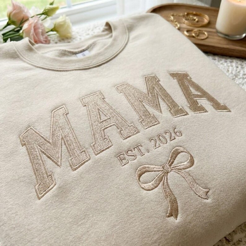 Peut inclure: Un sweat-shirt beige avec le mot "MAMA" brod&eacute; en police serif. En dessous, "EST. 2026" et un n&oelig;ud. Le sweat-shirt est sur une surface claire, avec des fleurs et des bijoux en arri&egrave;re-plan.