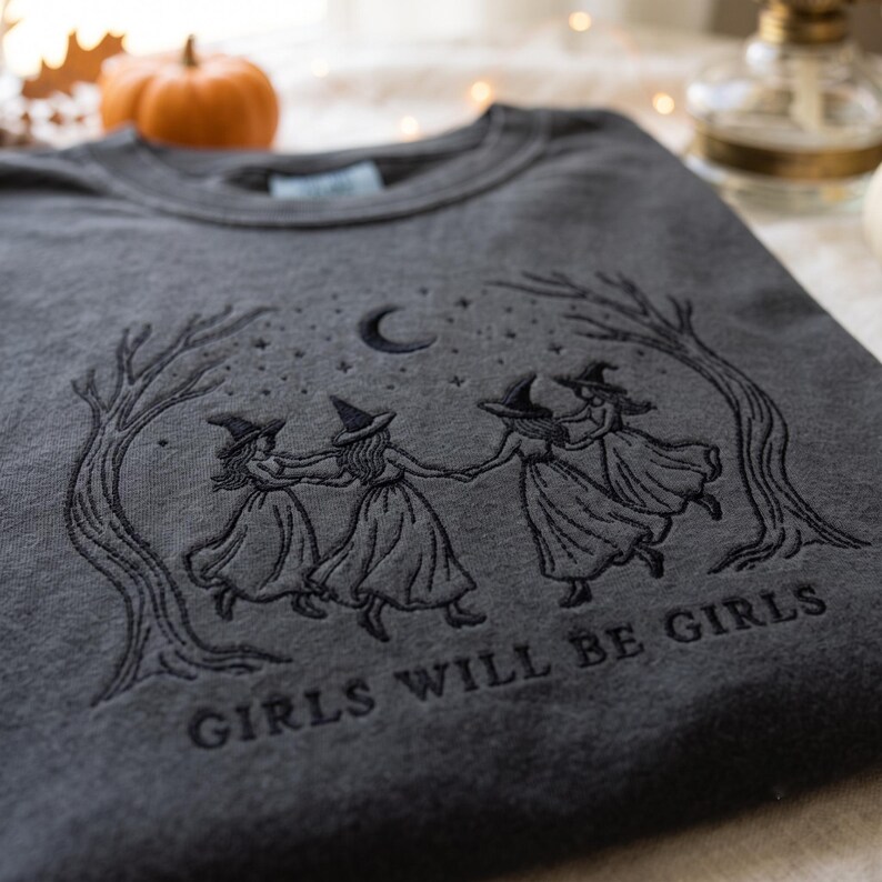 May include: A charcoal gray crewneck sweatshirt featuring black embroidery of four figures wearing witch hats and long dresses dancing in a circle between two trees under a crescent moon and stars, with the text GIRLS WILL BE GIRLS below.