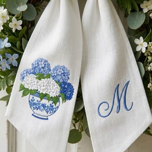 May include: Two white linen towels with blue embroidered details. One towel features a blue and white floral vase with blue and white hydrangeas. The other towel has a blue embroidered letter "M".