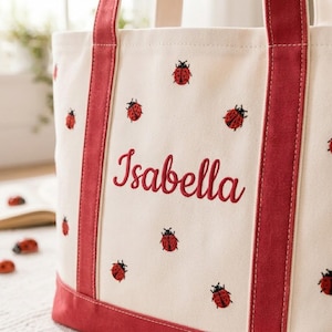 Pode incluir: Uma bolsa de lona cor de creme com alças e base de tecido vermelho. A bolsa é bordada com vários pequenos motivos de joaninhas vermelhas e pretas e o nome Isabella em uma fonte cursiva vermelha centralizada no painel frontal da bolsa.