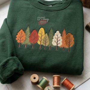 Könnte beinhalten: Ein dunkelgrüner Rundhals-Pullover mit einer gestickten Waldszene, die Bäume in Orange-, Rot-, Gelb- und Cremetönen zeigt. Der Text DThuy DECOR SINCE 2024 ist in Weiß über den Bäumen eingestickt. Darunter befinden sich vier hölzerne Garnrollen.