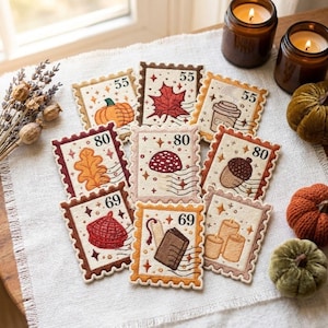 May include: A collection of nine rectangular embroidered patches designed like postage stamps. Each patch features autumn-themed motifs including a pumpkin, maple leaf, coffee cup, oak leaf, mushroom, acorn, beret, book, and candles with numbers.