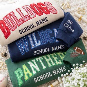 Puede incluir: Tres sudaderas dobladas en color crema, azul marino y verde oscuro. La sudadera crema presenta letras aplicadas rojas que dicen BULLDOGS, la azul marino dice WILDCATS y la verde dice PANTHERS. Cada una incluye el texto SCHOOL NAME.