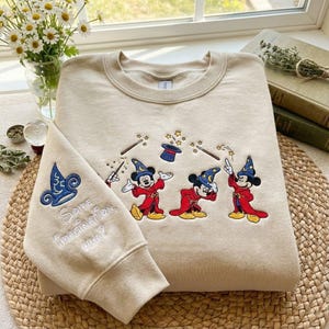 May include: Beige sweatshirt with embroidered Mickey Mouse characters in wizard attire. The design includes wands, stars, and a hat. The left sleeve features a blue and white design with the text "Some Imagination, Huh?"