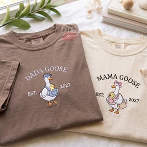 May include: A brown t-shirt with a white embroidered goose wearing a blue cap and carrying a yellow chick in a carrier, labeled DADA GOOSE EST. 2027, and a cream t-shirt with a white goose wearing a pink bow, labeled MAMA GOOSE EST. 2027.