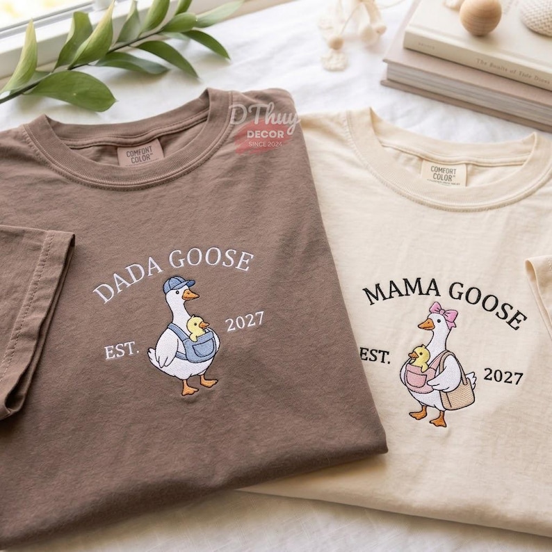 May include: A brown t-shirt with a white embroidered goose wearing a blue cap and carrying a yellow chick in a carrier, labeled DADA GOOSE EST. 2027, and a cream t-shirt with a white goose wearing a pink bow, labeled MAMA GOOSE EST. 2027.