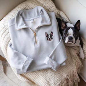 May include: A light gray sweatshirt with a gold zipper and a Boston Terrier dog patch. The cuffs are embroidered with the name "Noah". A Boston Terrier dog is next to the sweatshirt.