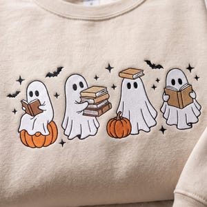 May include: A beige crewneck sweatshirt featuring an embroidered design of four white ghosts. The ghosts are depicted interacting with orange pumpkins, brown books, and small black bats and stars scattered across the fabric surface.
