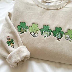 May include: Beige sweatshirt featuring embroidered green shamrocks, each with a different item: a book, pencil, map, globe, and paper. A pencil with a green bow is embroidered on the sleeve. The sweatshirt has a crew neck and cuffed sleeves.