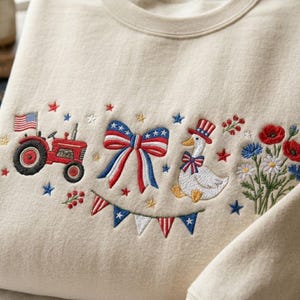 K&ouml;nnte beinhalten: Cremefarbenes Sweatshirt mit gestickten patriotischen Designs. Mit einem roten Traktor mit amerikanischer Flagge, einer rot-wei&szlig;-blauen Schleife, einer Gans mit Zylinder und floralen Akzenten. Enth&auml;lt ein Banner aus rot-wei&szlig;-blauen Wimpeln.
