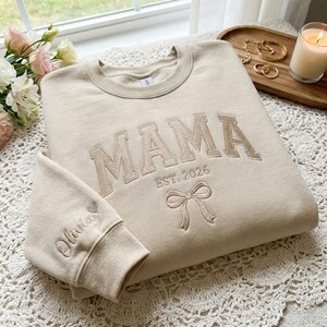 Peut inclure: Sweat-shirt beige avec le mot "MAMA" brod&eacute; en beige, ainsi que "EST. 2026" et un n&oelig;ud. Le poignet de la manche porte le nom "Olivia" et un c&oelig;ur. Le sweat-shirt est pos&eacute; sur une surface en dentelle blanche.