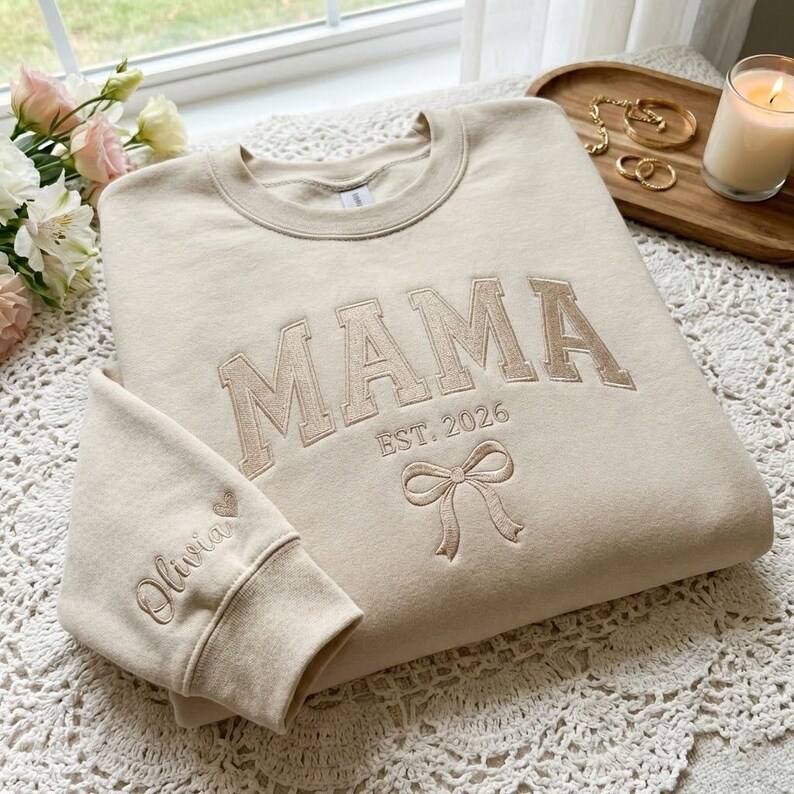 Peut inclure: Sweat-shirt beige avec le mot "MAMA" brod&eacute; en beige, ainsi que "EST. 2026" et un n&oelig;ud. Le poignet de la manche porte le nom "Olivia" et un c&oelig;ur. Le sweat-shirt est pos&eacute; sur une surface en dentelle blanche.