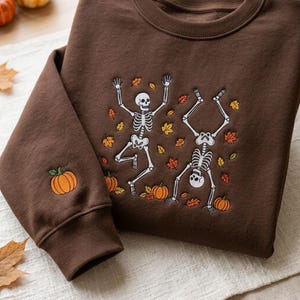 Op de afbeelding: Een donkerbruine sweater met ronde hals voorzien van een borduursel van twee witte skeletten die dansen tussen verspreide oranje en gele herfstbladeren en kleine pompoenen. Op de mouw is een extra kleine oranje pompoen geborduurd.