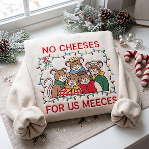 Può includere: Una felpa color crema con un ricamo raffigurante cinque topi che indossano abiti invernali. Il testo "NO CHEESES FOR US MEECES" è cucito in rosso sopra e sotto i topi, il tutto incorniciato da una catena di luci natalizie colorate.