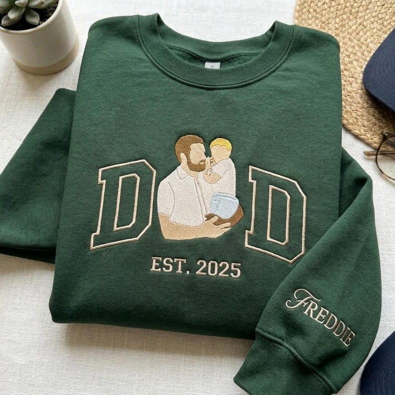 May include: A dark green sweatshirt with the word "DAD" in white block letters and an embroidered image of a father holding a child. The text "EST. 2025" is below the image, and the name "Freddie" is embroidered on the sleeve.