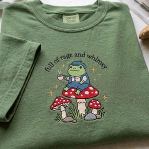 May include: An olive green t-shirt featuring an embroidered design of a green frog wearing a blue outfit, holding a white mug, and sitting on a red mushroom. The text "full of rage and whimsy" is arched above the frog with small gold stars.