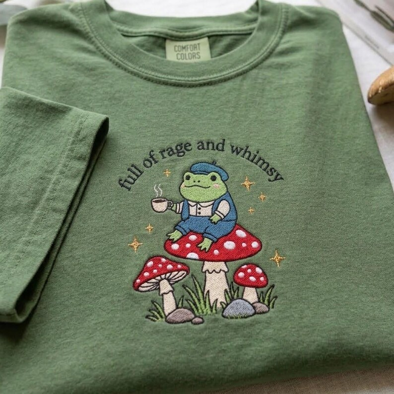 May include: An olive green t-shirt featuring an embroidered design of a green frog wearing a blue outfit, holding a white mug, and sitting on a red mushroom. The text "full of rage and whimsy" is arched above the frog with small gold stars.