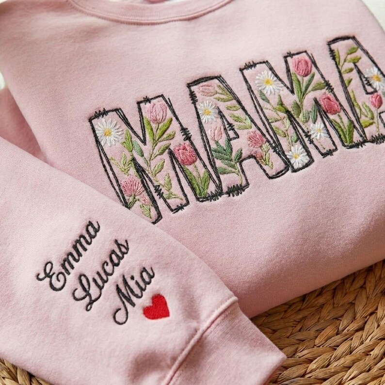 Op de afbeelding: Een lichtroze sweatshirt met het woord "MAMA" geborduurd in grote letters gevuld met bloemmotieven. De mouw heeft de namen "Emma", "Lucas" en "Mia" geborduurd in zwart, met een rood hartje eronder.