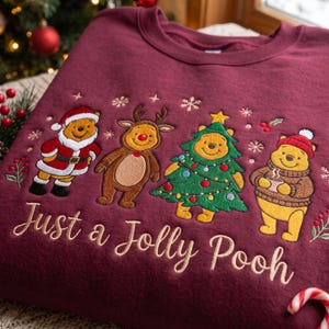 Può includere: Una felpa girocollo color bordeaux con illustrazioni ricamate dei personaggi di Winnie the Pooh in abiti festivi, tra cui un costume da Babbo Natale, corna di renna, un costume da albero di Natale e un maglione invernale, con il testo Just a Jolly Pooh.