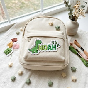 May include: A cream-colored canvas backpack featuring a green embroidered dinosaur patch and the name NOAH in green letters on the front pocket. The backpack is surrounded by small green and cream paper stars and colorful embroidery floss.