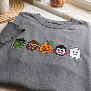 May include: A charcoal gray t-shirt featuring five embroidered character heads in a row. The designs include a green monster, a brown werewolf, an orange jack-o'-lantern, a vampire with red accents, and a plain white head with a simple smile.