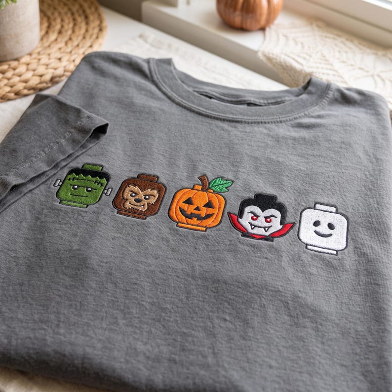 May include: A charcoal gray t-shirt featuring five embroidered character heads in a row. The designs include a green monster, a brown werewolf, an orange jack-o'-lantern, a vampire with red accents, and a plain white head with a simple smile.