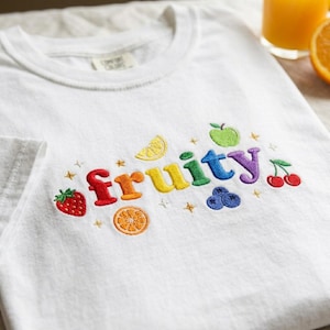 May include: A white t-shirt featuring colorful embroidered text that reads fruity, surrounded by small embroidered fruit icons including a strawberry, lemon slice, orange slice, green apple, blueberries, and cherries, along with small gold star accents.