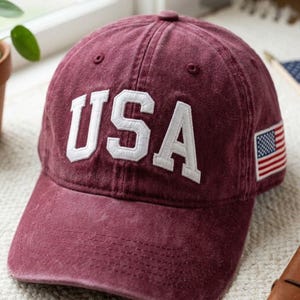 May include: Maroon baseball cap with the word "USA" in white, embroidered letters. The cap features a small American flag patch on the side. The hat is made of a soft, worn material.
