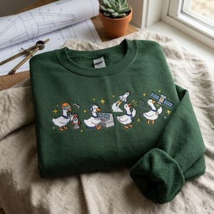 May include: A dark green sweatshirt with an embroidered design of cartoon geese in engineering roles. The geese are shown with tools, computers, and a rocket. The design includes yellow stars and is on a crew neck sweatshirt.