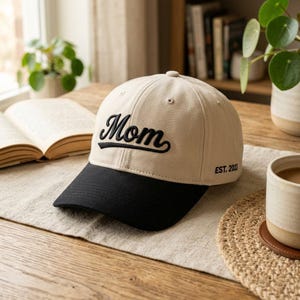 May include: A beige baseball cap with a black brim. The word "Mom" is embroidered in black script on the front, with "EST. 2023" below. The hat is on a wooden surface.