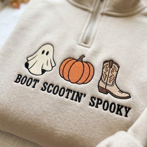 May include: A beige quarter-zip sweatshirt featuring embroidered designs of a white ghost, an orange pumpkin, and a tan cowboy boot. Below the graphics, the text BOOT SCOOTIN' SPOOKY is embroidered in black capital letters across the chest area.