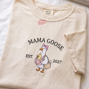 May include: A cream-colored short-sleeve t-shirt featuring an embroidered graphic of a white goose wearing a pink bow, carrying a yellow duckling in a pink carrier and a tan bag. The text reads MAMA GOOSE EST. 2027 and COMFORT COLOR D THUY DECOR.