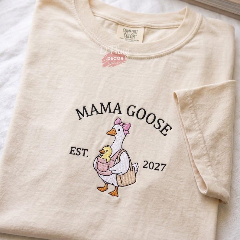 May include: A cream-colored short-sleeve t-shirt featuring an embroidered graphic of a white goose wearing a pink bow, carrying a yellow duckling in a pink carrier and a tan bag. The text reads MAMA GOOSE EST. 2027 and COMFORT COLOR D THUY DECOR.